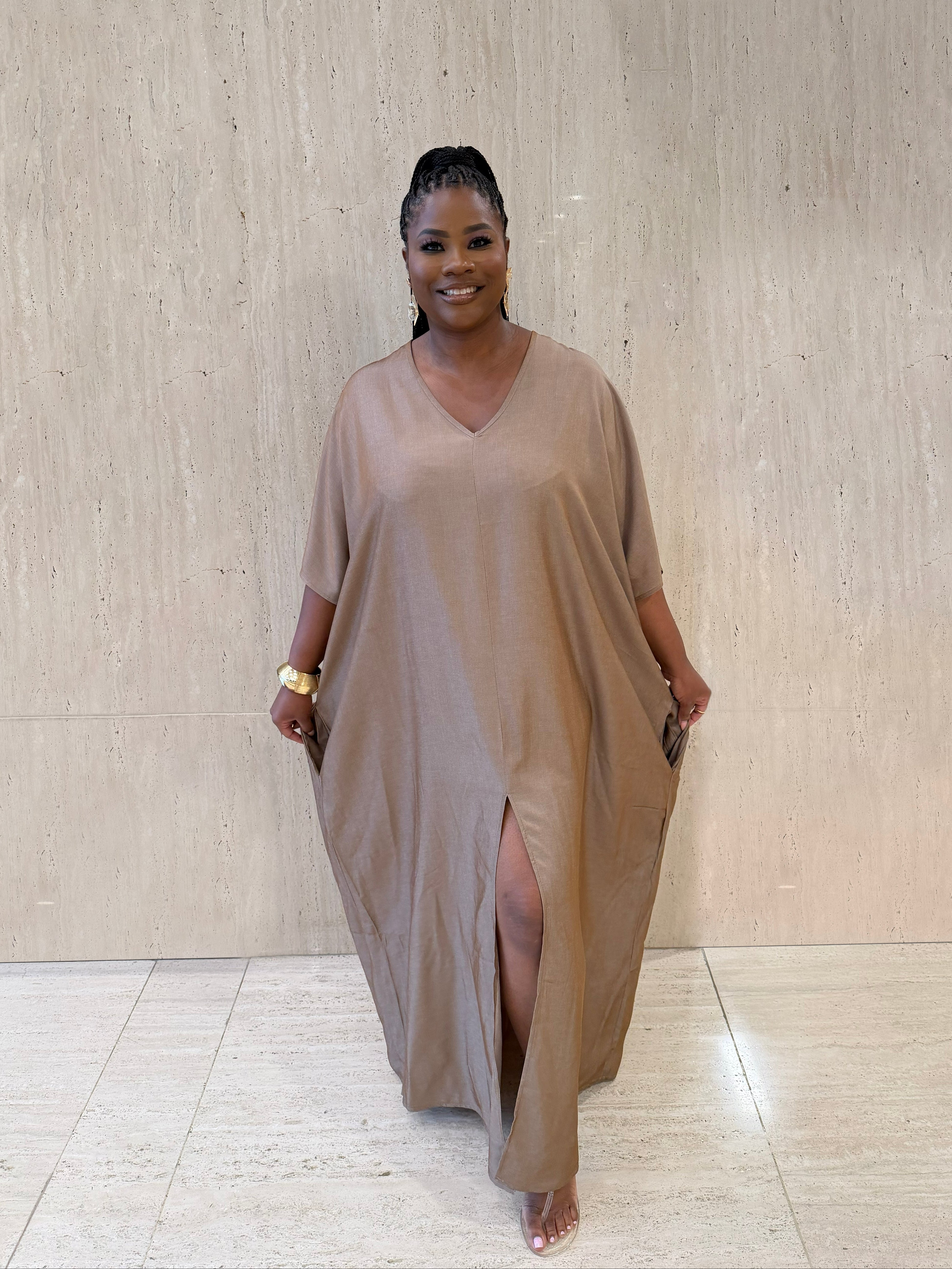 Woman wearing a long, light brown dress standing against a beige wall.