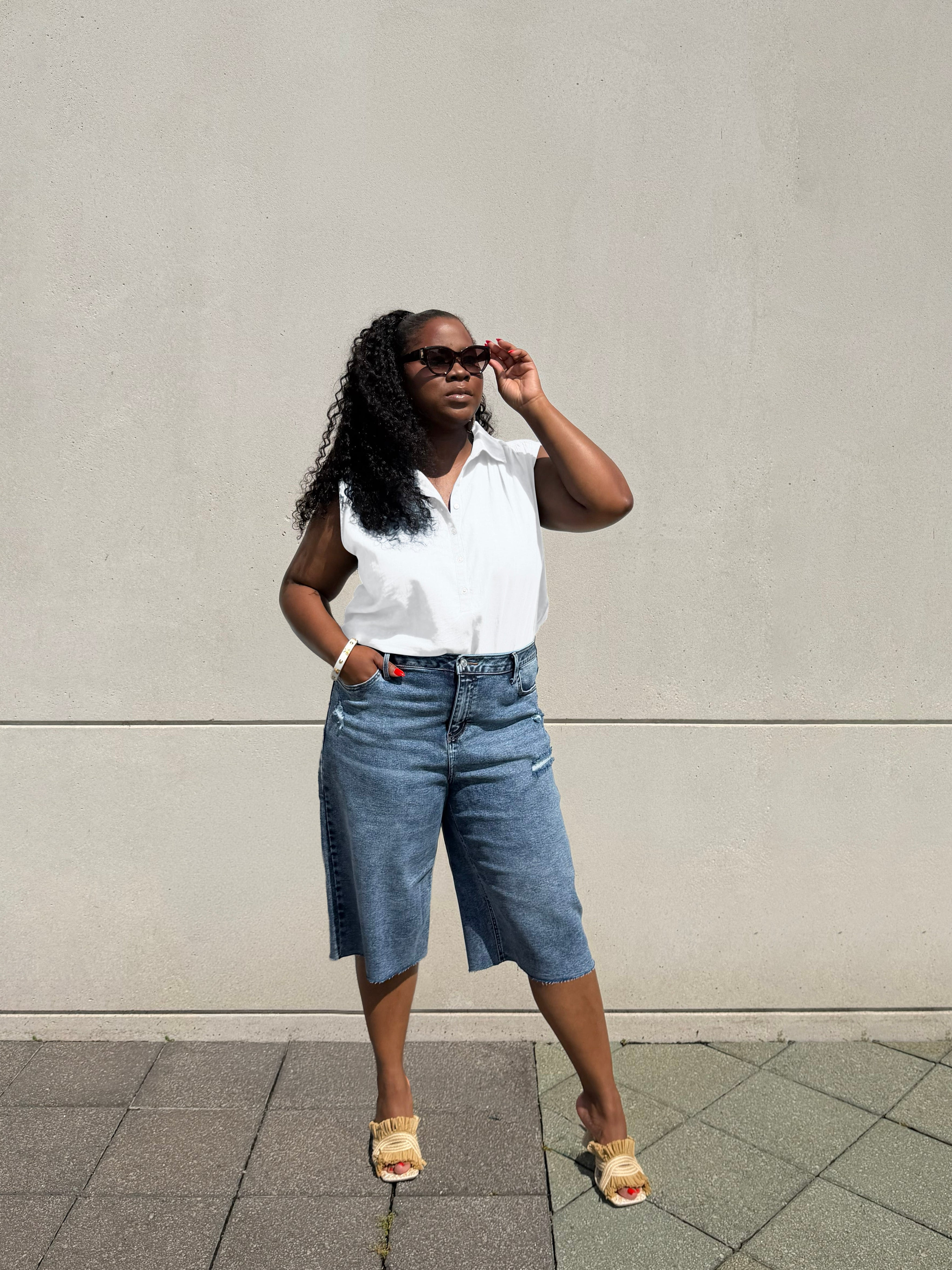 A woman posing outdoors against a concrete wall, wearing a white sleeveless button-up shirt, blue denim cropped shorts, and beige sandals.