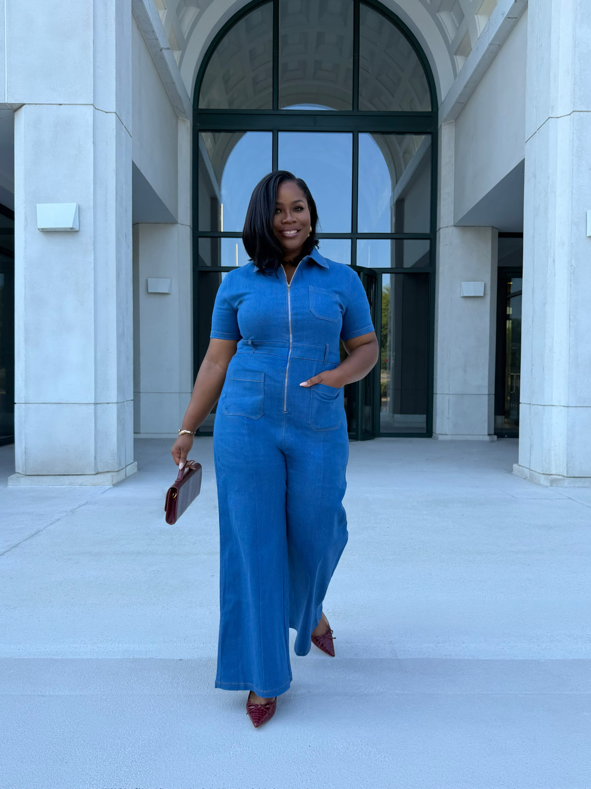 A woman wearing a blue denim short-sleeve jumpsuit with a front zipper, walking outdoors in front of a modern building with large arched windows and columns.