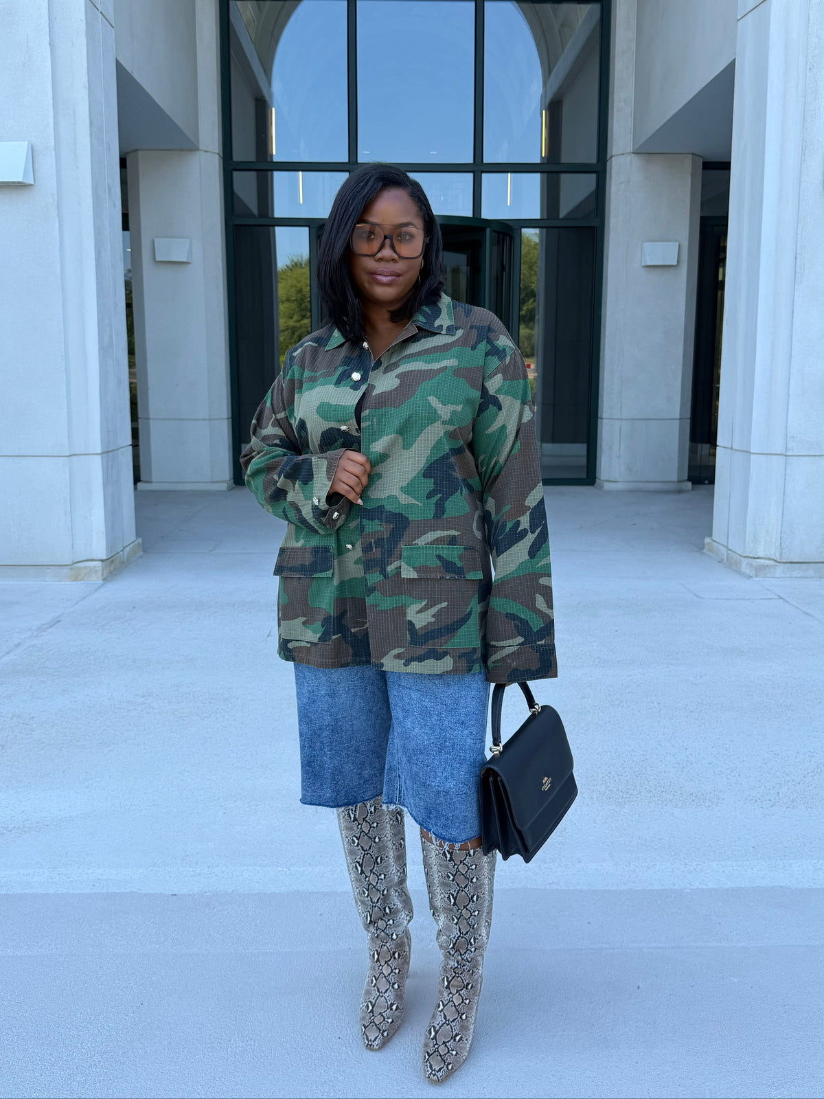 A woman stands in front of a modern building entrance wearing a camouflage button-up jacket, a denim jorts, snake skin knee-high boots, and holding a black handbag.