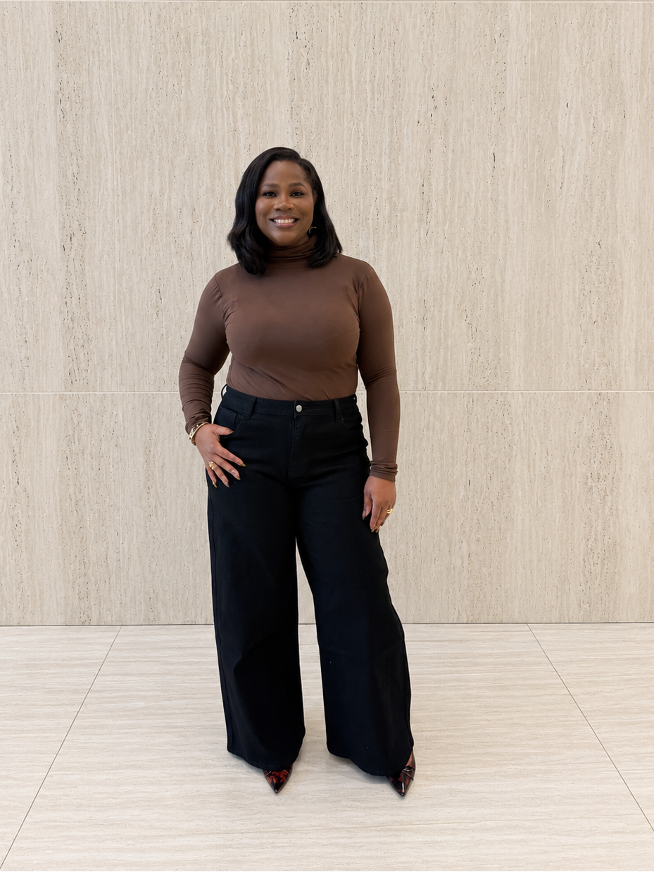 A woman stands in front of a light beige textured wall wearing a fitted brown long-sleeve top and black wide-leg pants.