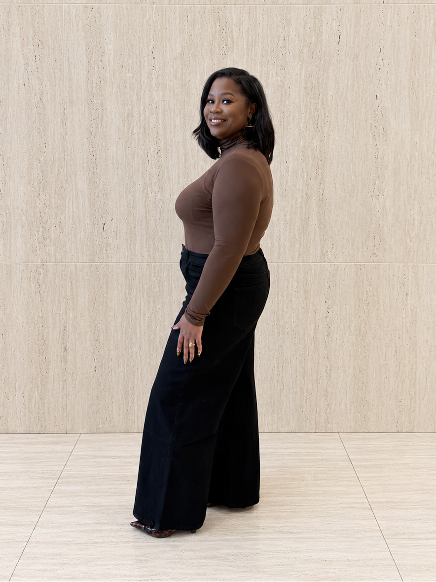 A woman stands in profile against a light beige textured wall and tiled floor, wearing a fitted brown long-sleeve top and black wide-leg pants.