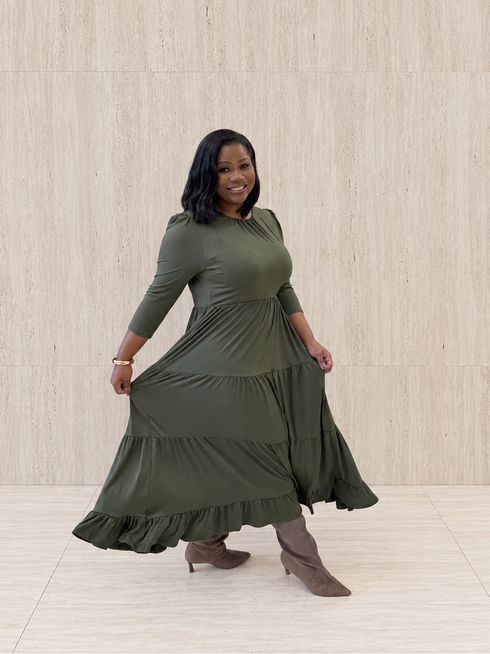 A woman models a three-quarter-sleeve olive-green tiered maxi dress with ruffled trim, paired with taupe heeled boots, standing against a light beige, wood-paneled wall.