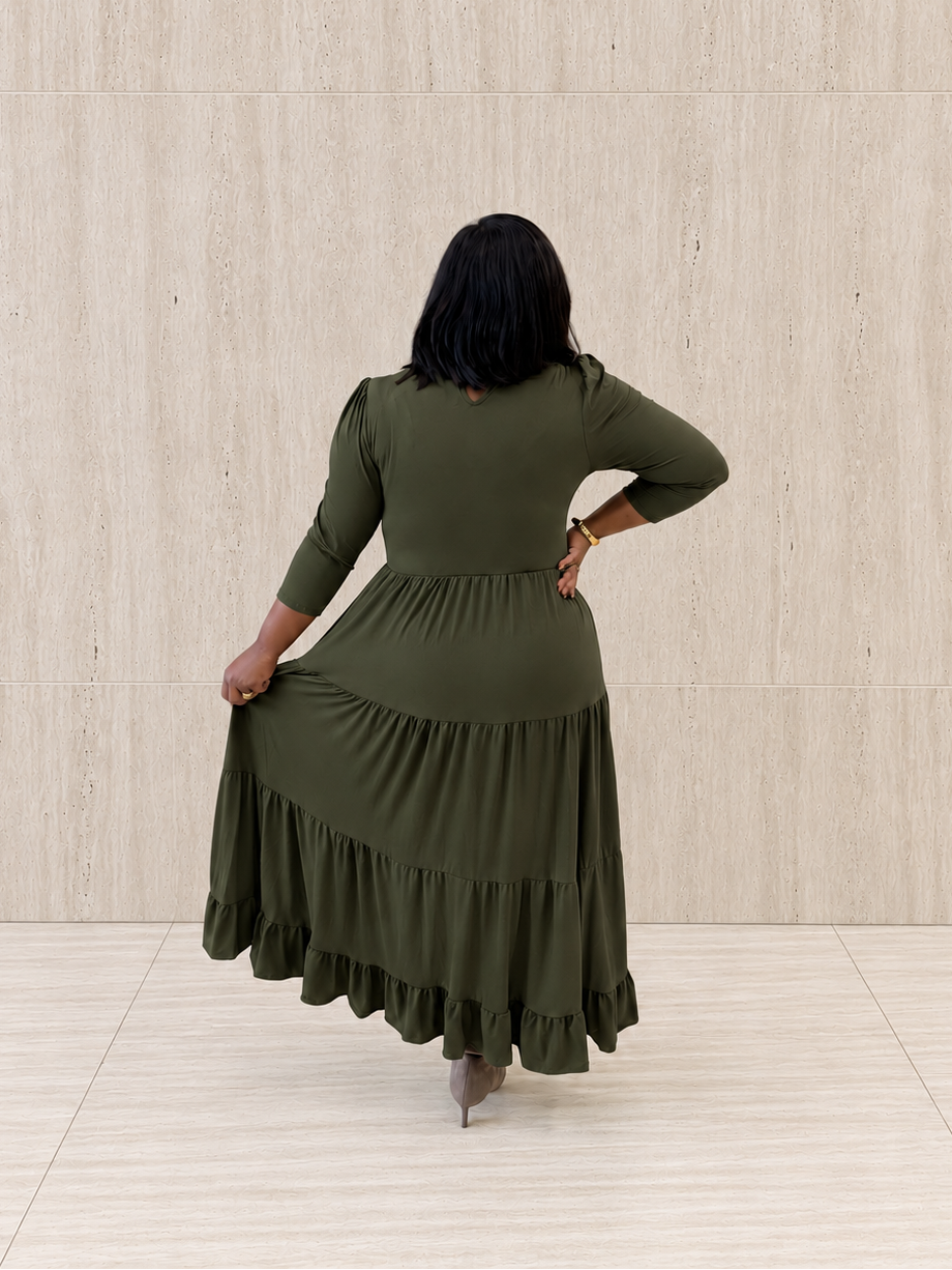A woman viewed from behind wearing an olive green tiered maxi dress with three-quarter sleeves, standing against a light beige textured wall and tiled floor.