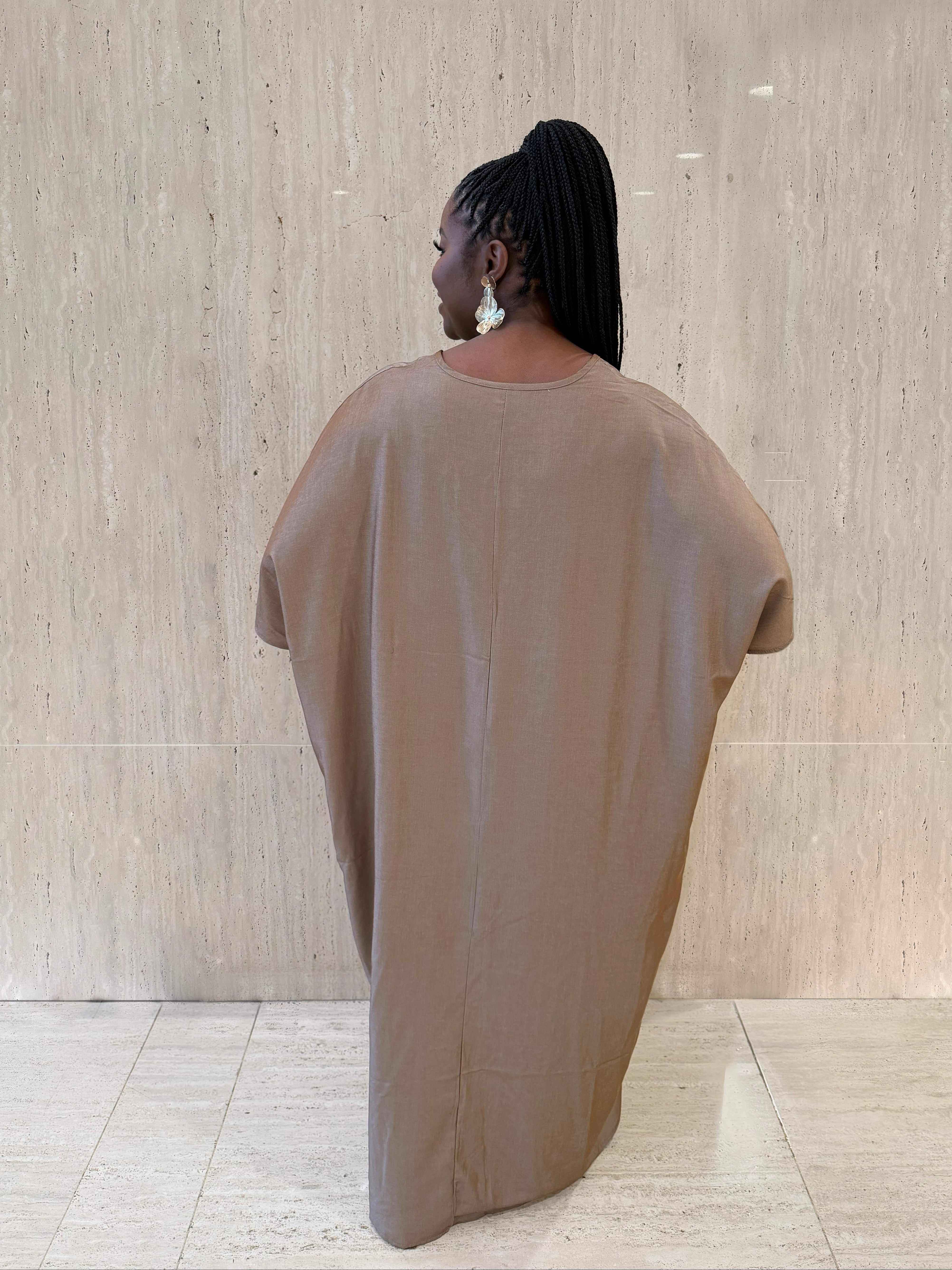 Person wearing a beige oversized garment against a light wooden paneled wall.
