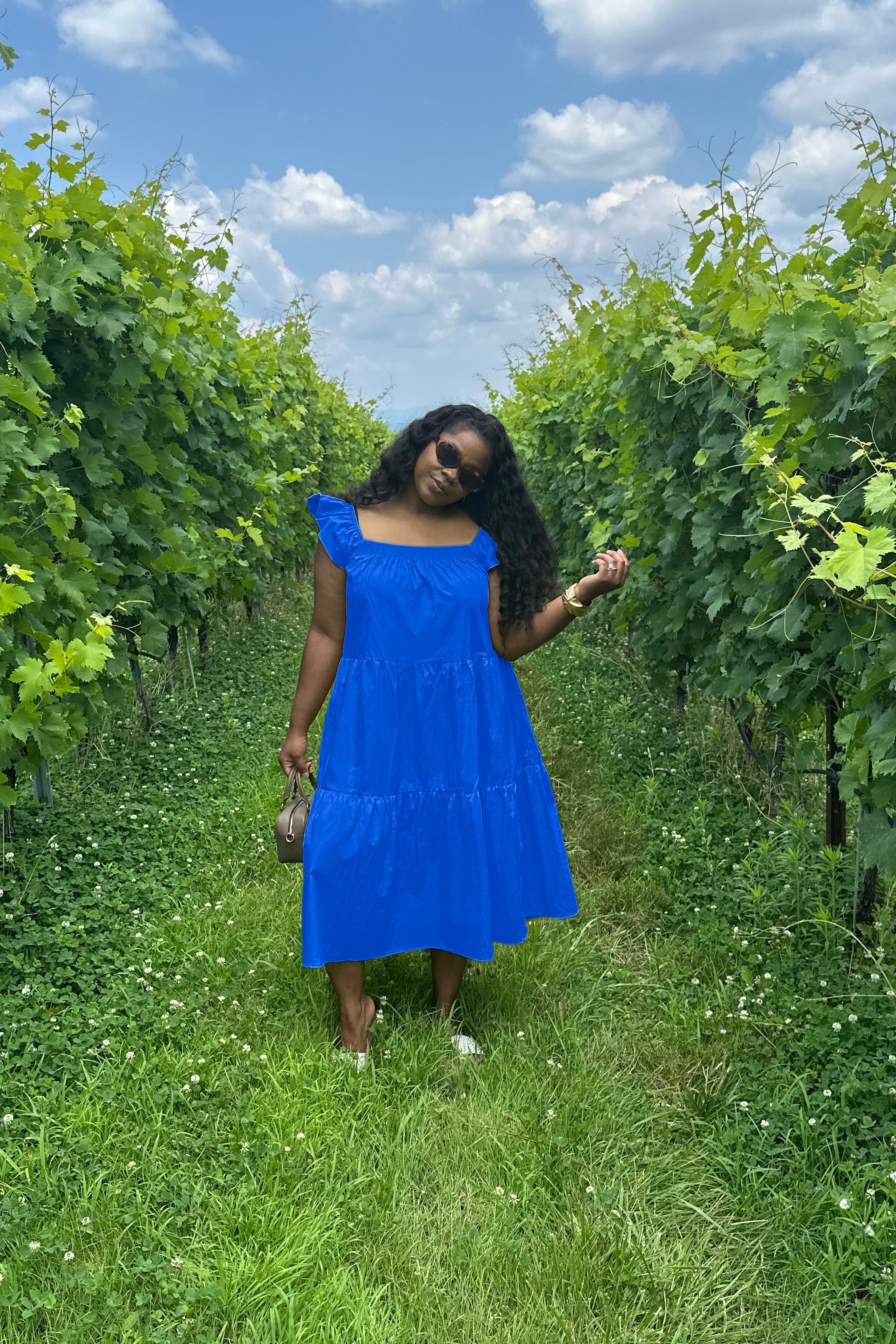 Woman in a blue dress standing in a vineyard with green leaves and blue sky.