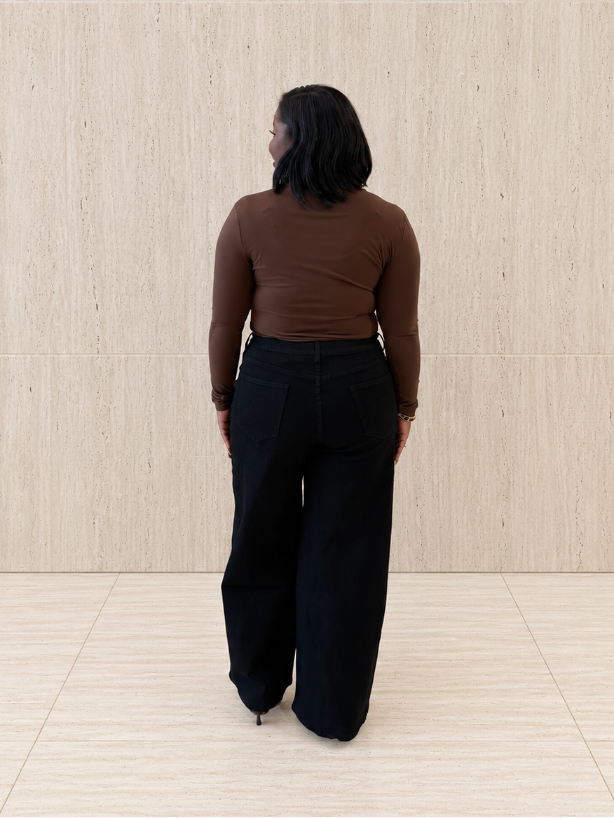 A woman viewed from behind wearing a brown long-sleeve top and black wide-leg pants, standing against a light beige wood-paneled wall and floor.