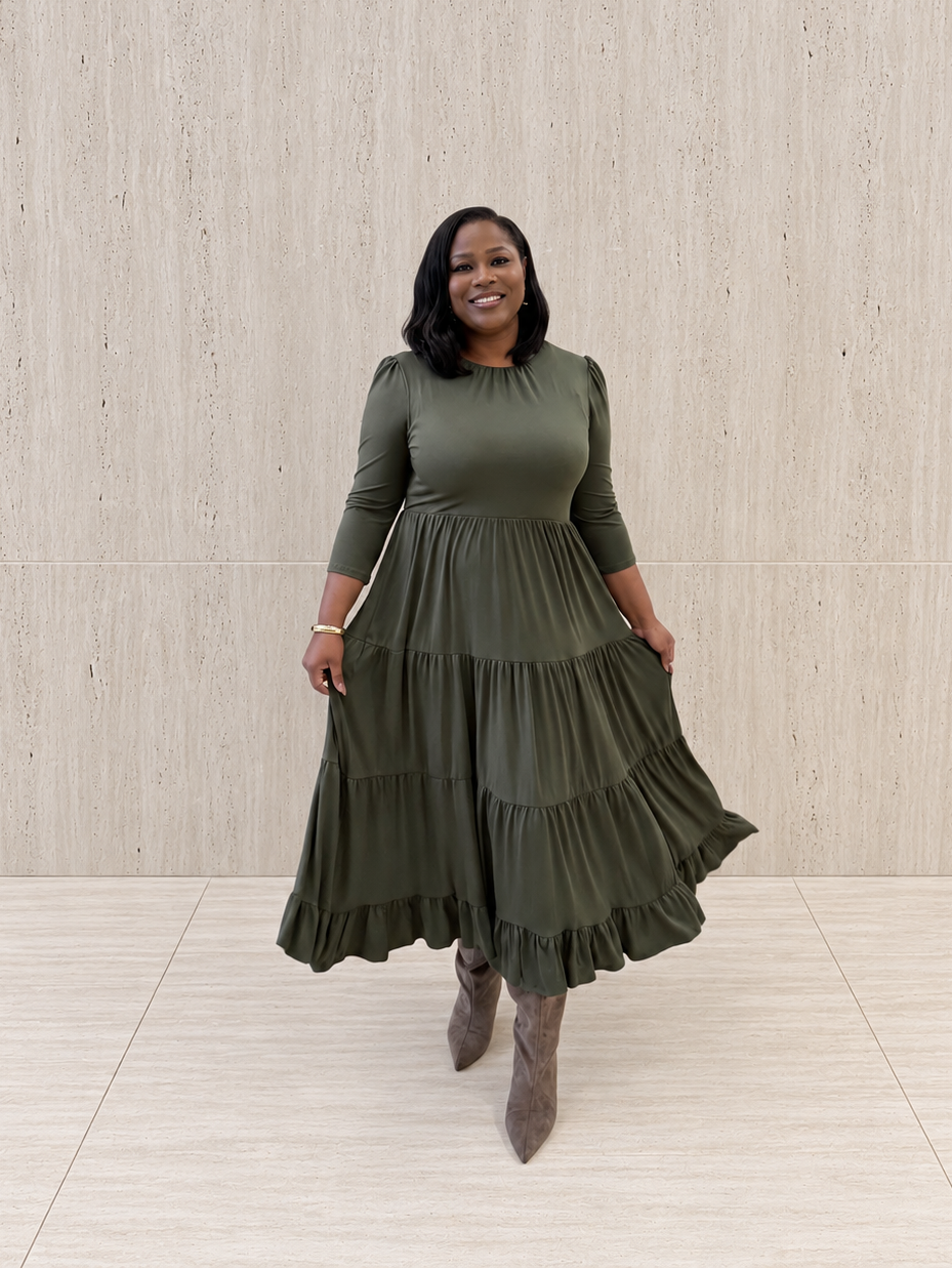 A woman in a three-quarter-sleeve olive-green tiered maxi dress and taupe pointed-toe boots stands against a light beige textured wall.