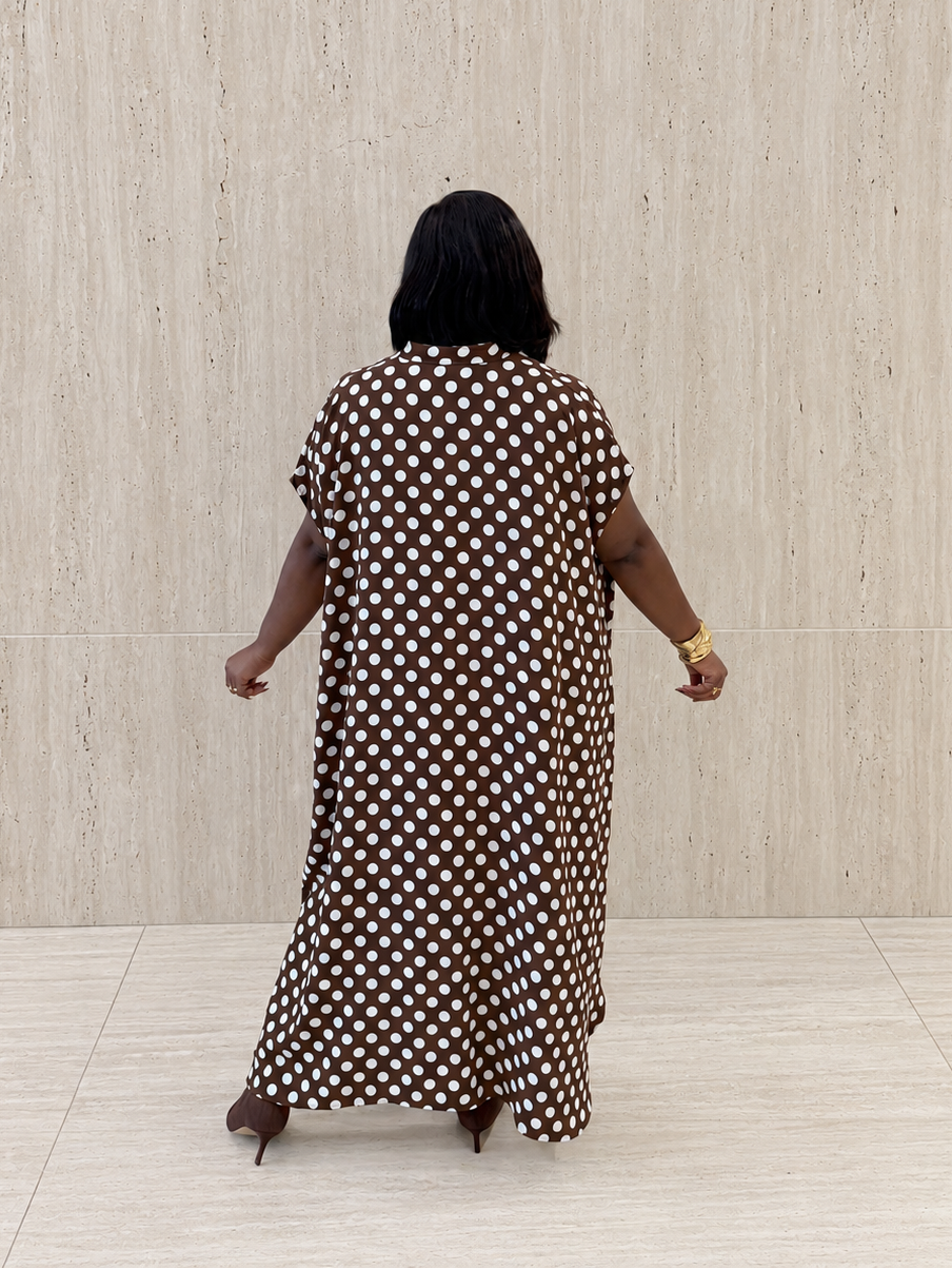 A woman viewed from behind wearing a brown and white polka dot maxi dress, standing against a light beige textured wall and tiled floor.