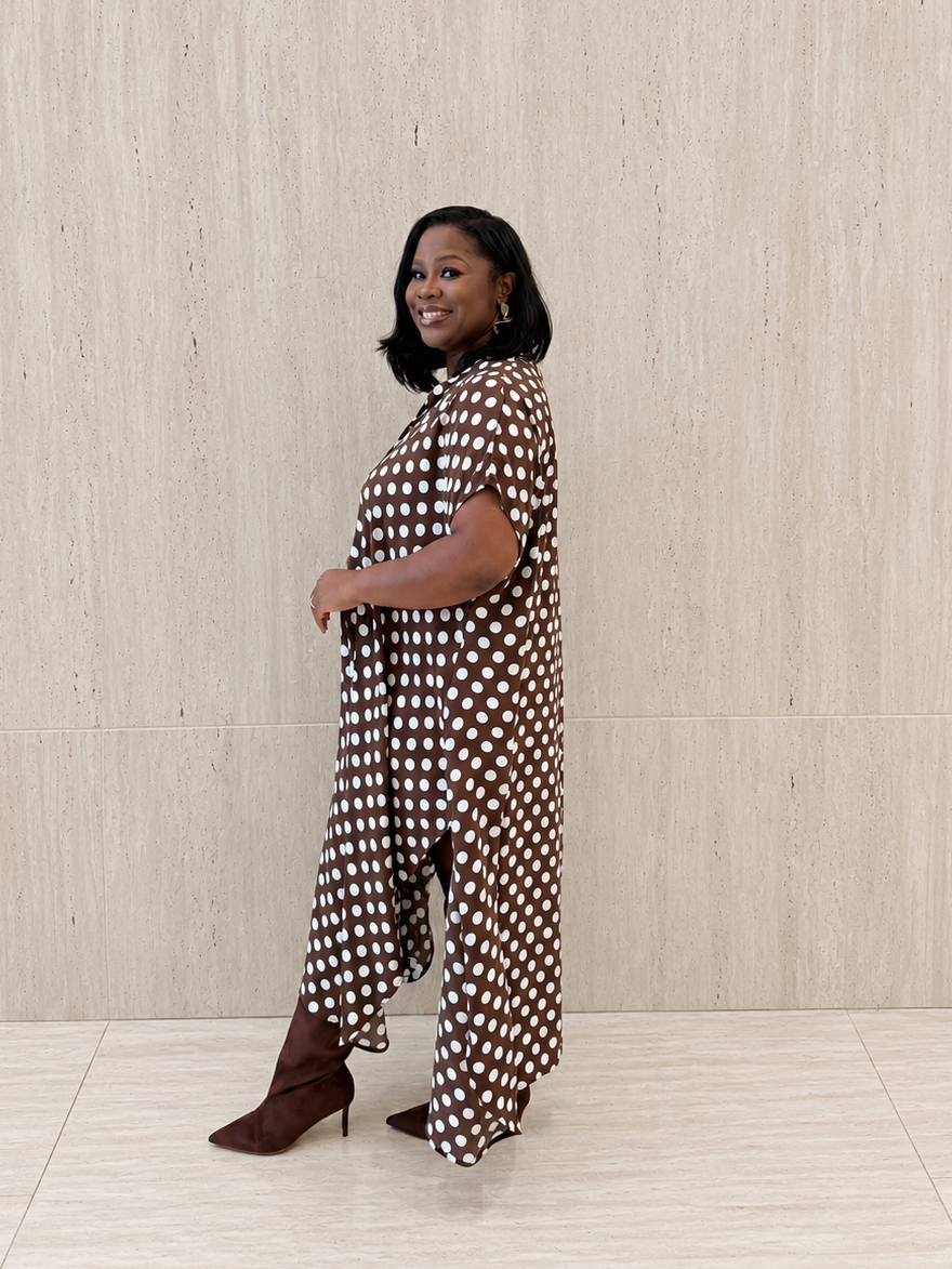 A woman modeling a brown and white polka dot jumpsuit with wide-leg pants and short sleeves, paired with brown heeled boots, standing against a light beige textured wall.