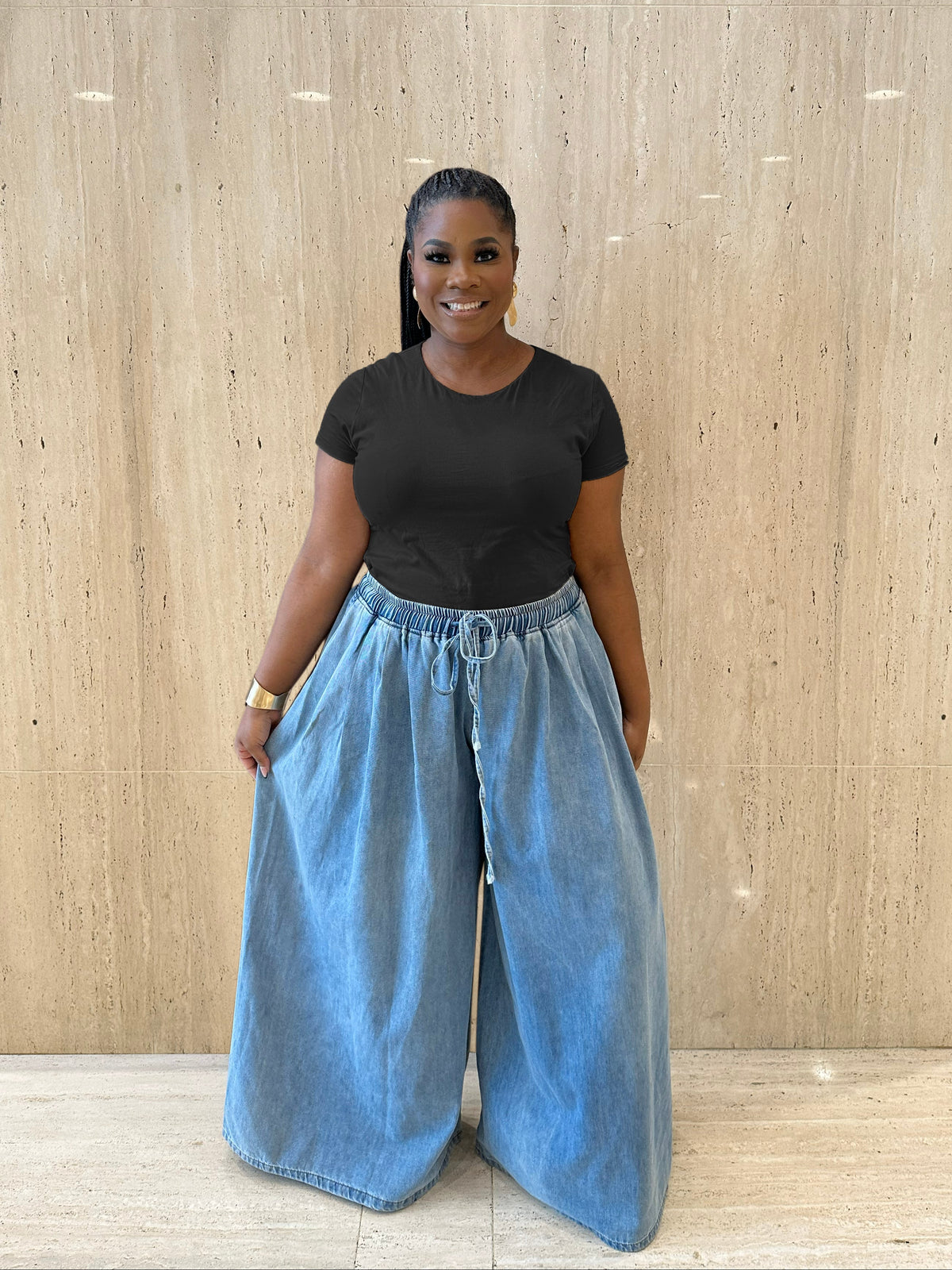 A woman stands in front of a beige stone wall wearing a black short-sleeve t-shirt and light blue wide-leg denim pants.