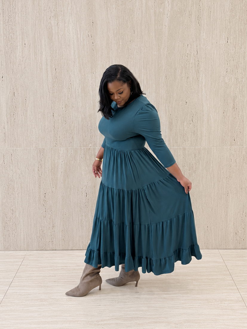 A woman wearing a teal blue tiered maxi dress with three-quarter sleeves and taupe ankle boots, standing against a light beige textured wall.