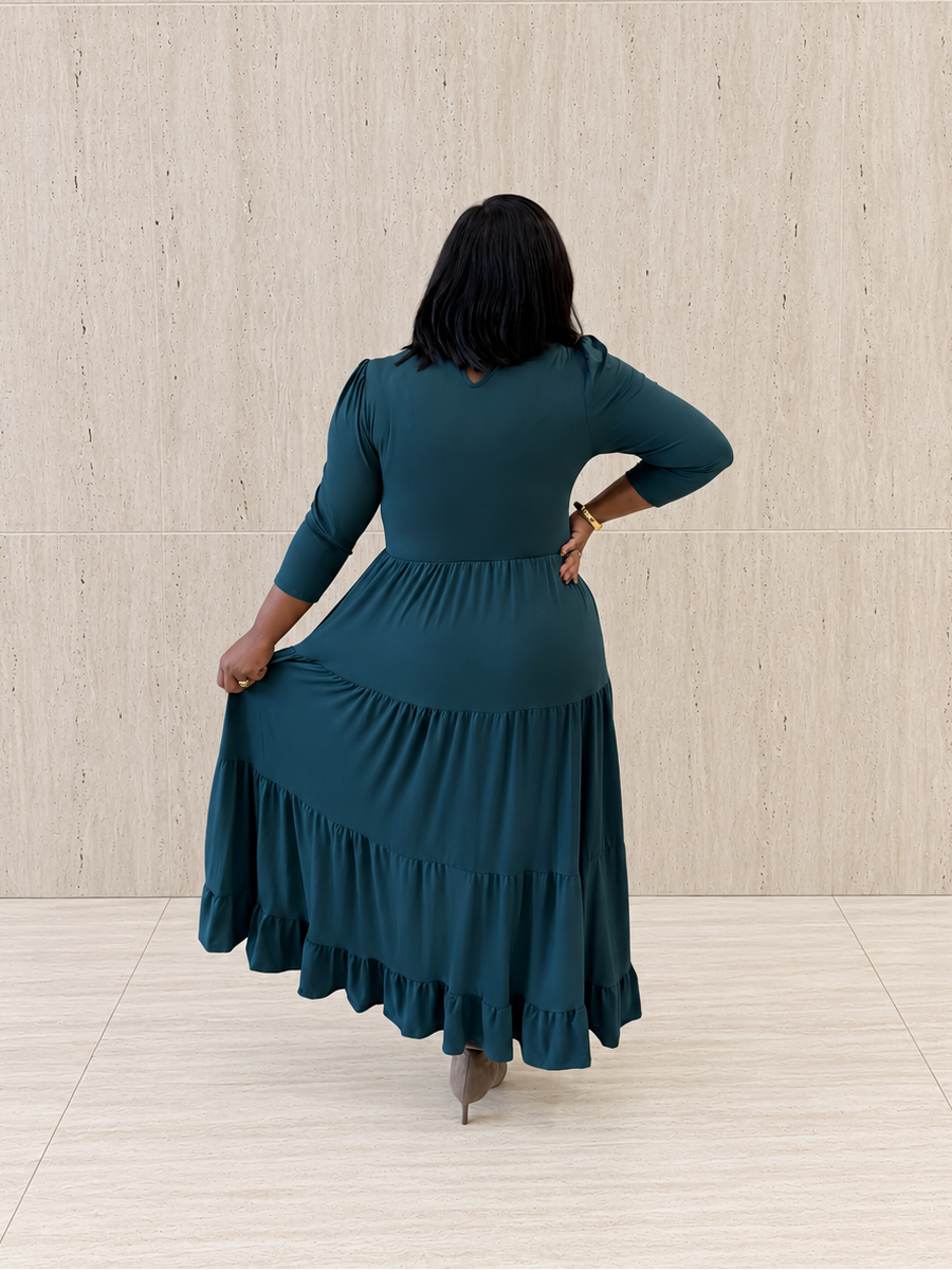 A woman wearing a dark teal green tiered maxi dress, shown from the back as she holds up the skirt to show its volume. She is standing in front of a light beige textured wall and tiled floor.