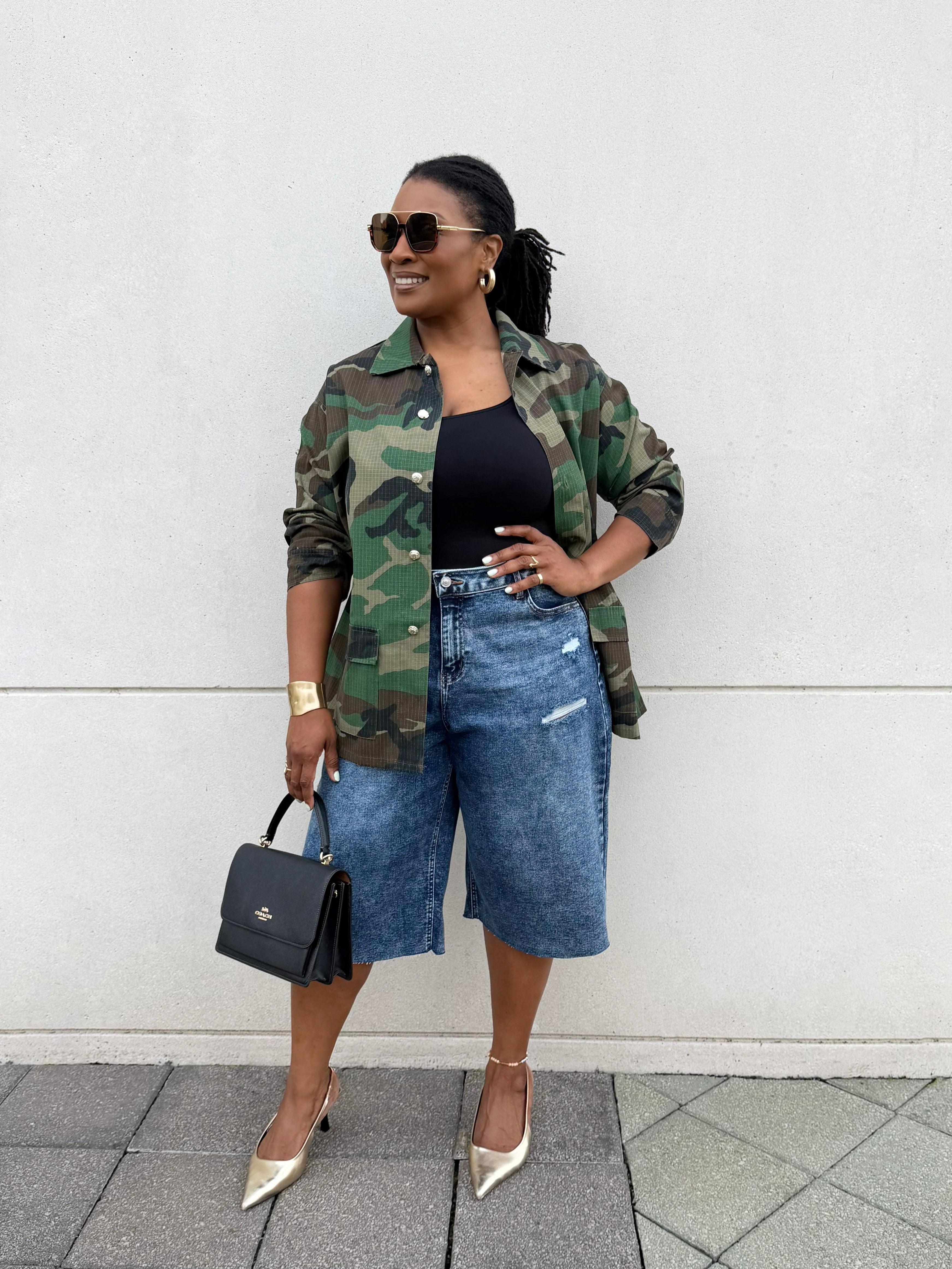 A woman stands against a white wall wearing a green camouflage button-up jacket over a black top, paired with high-waisted blue denim shorts and beige pointed-toe heels. She is holding a small black handbag.