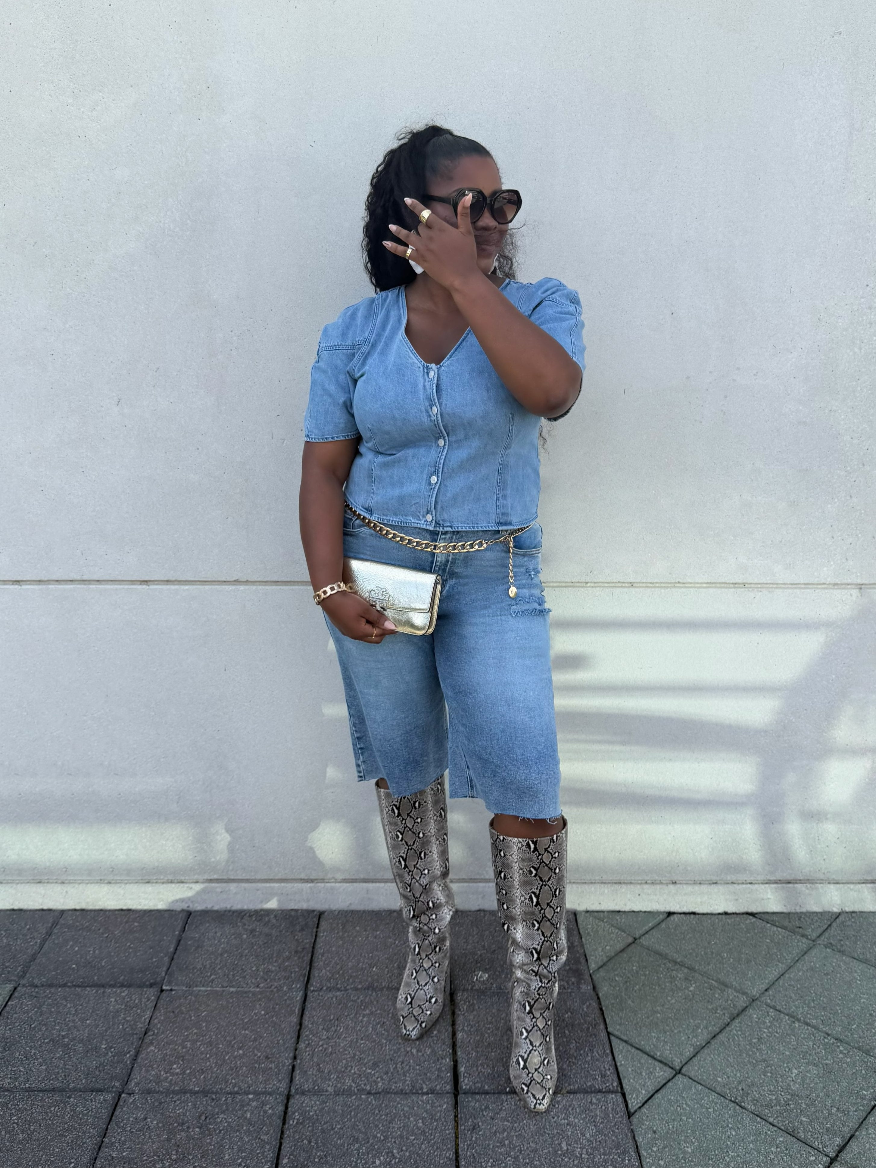 A woman posing outdoors against a light gray concrete wall, wearing a light blue denim short-sleeve top and matching denim jorts, styled with a gold chain belt, a gold clutch, and snakeskin-patterned knee-high boots.